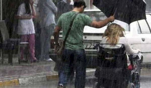 In rain man walks beside woman in wheelchair holding umbrella over her