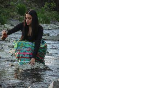 Autumn Peltier wearing a black top and multi-coloured skirt, squatting in a river and pouring water from a mug